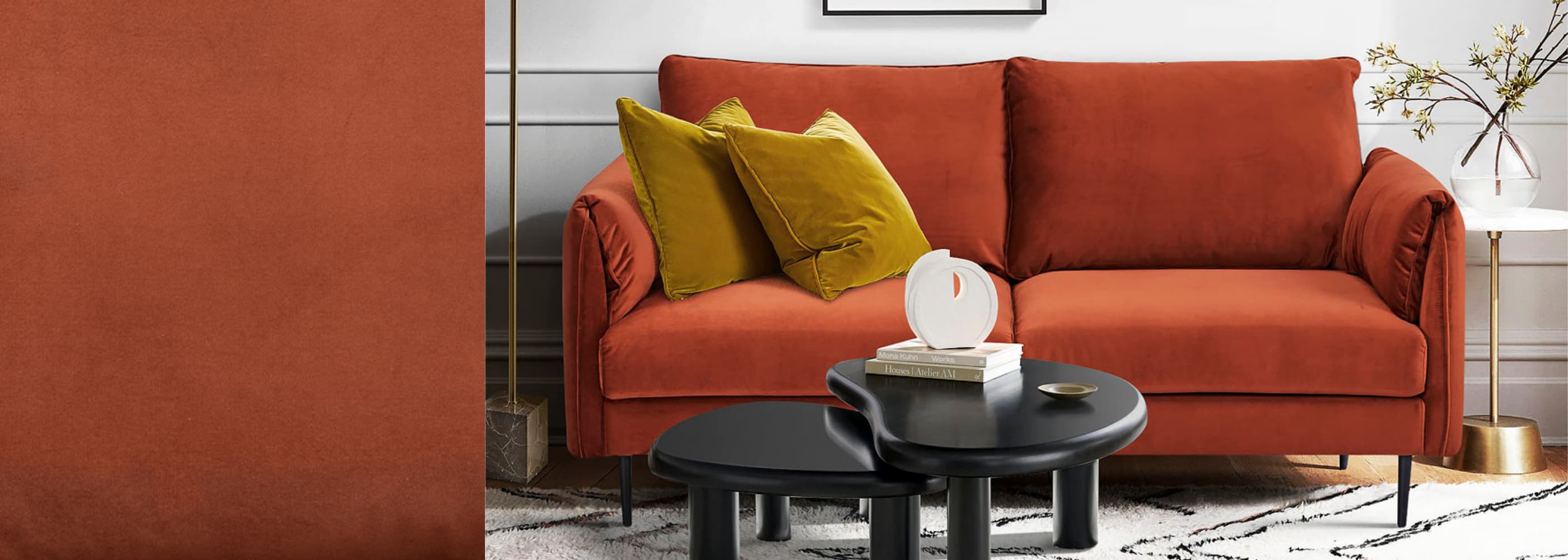 Canapé 2 places en velours terracotta avec accoudoirs arrondis et pieds fins noirs, décoré de coussins jaune moutarde, dans un salon moderne avec tables basses noires et éléments dorés.